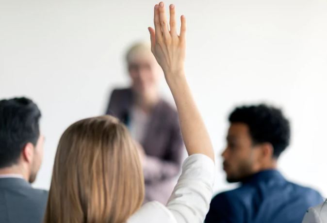 a stock image of a student with long blonde hair raising their hand, other students are blurred in the background 