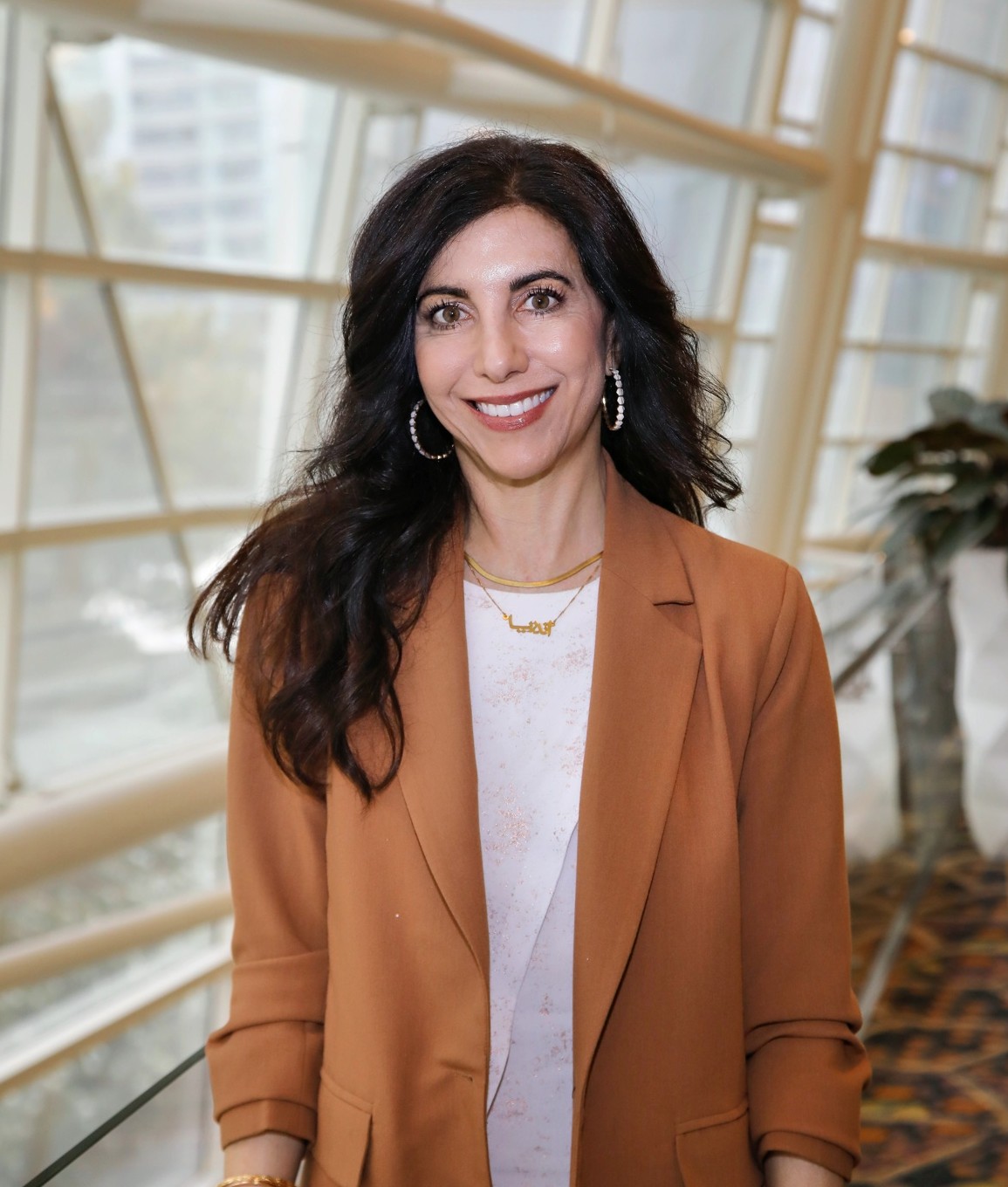 a photo of Andrea Shaheen, a woman with long black hair and a white shirt and an a orange colored blazer and gold jewelry
