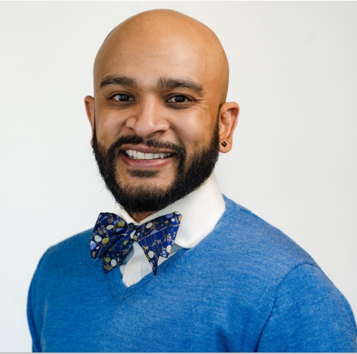 head shot of Mitesh Patel, a man with brown skin, a beard, a shaved head, he is wearing earing in his left lobe a white collared short a blue bow tie and a blue sweater
