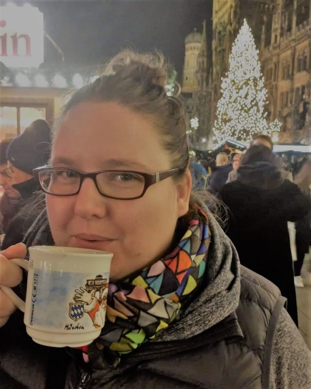 photo of Sarah Brockmeyer drinking gluhwein at a Christmas market in Munich Germany her brown hair is in a bun, she is wearing a multicolored scarf and glasses